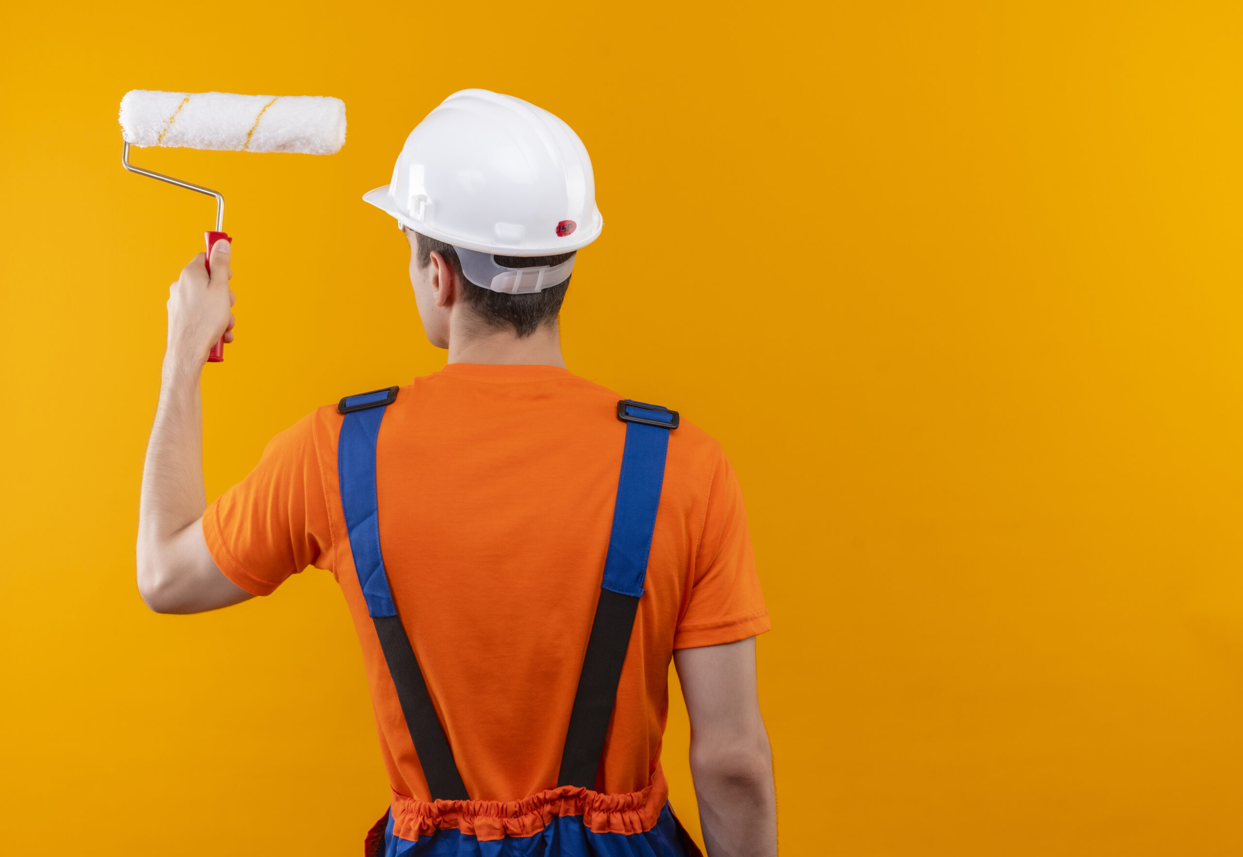 young builder man wearing construction uniform and white safety helmet paints the wall with roller brush over orange isolated background